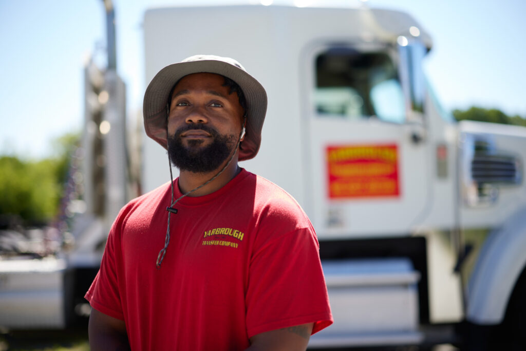 Man wearing a red shirt and gray bucket hat stands in front of a white truck with a red and yellow logo on the door, outdoors on a sunny day.