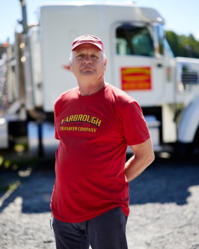 An older man wearing a red cap and red t-shirt stands in front of a white truck. The t-shirt and cap both display Yarbrough Transfer Company in yellow. The man has his hands behind his back, and the ground is gravel.